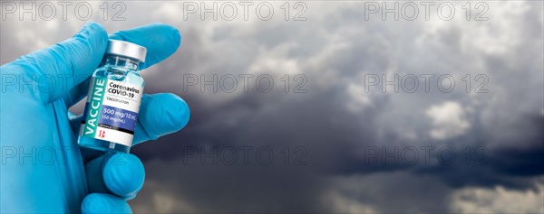 Doctor or nurse holding coronavirus COVID-19 vaccine vial against stormy clouds background banner