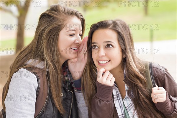 Two attractive mixed-race woman with backpacks whispering secrets outside