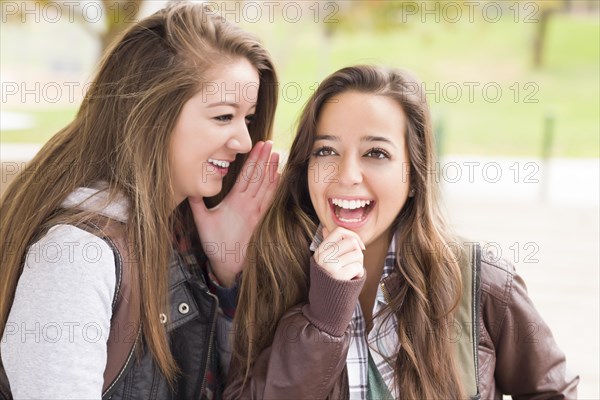 Two attractive mixed-race woman with backpacks whispering secrets outside