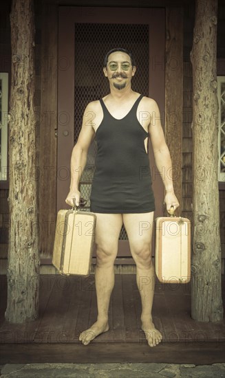 Goofy gentleman dressed in 1920's era swimsuit holding suitcases on porch of cabin