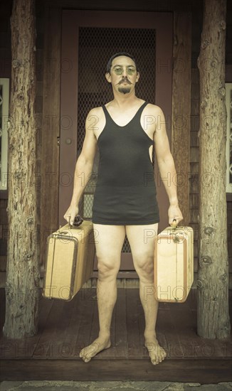 Goofy gentleman dressed in 1920's era swimsuit holding suitcases on porch of cabin