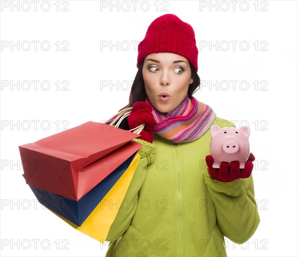 Concerned mixed-race woman wearing winter clothes holding shopping bags and piggybank isolated on white background