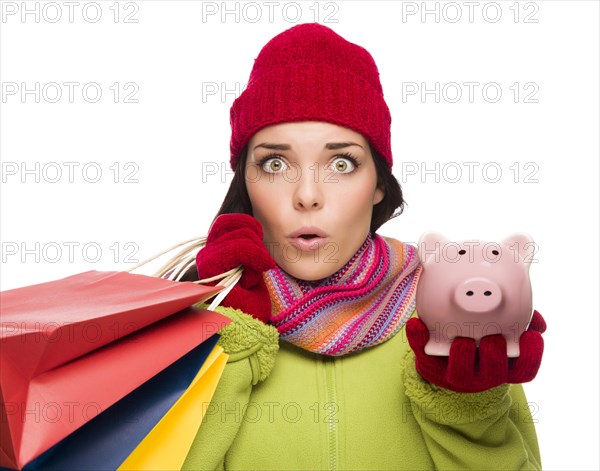 Concerned mixed-race woman wearing winter clothes holding shopping bags and piggybank isolated on white background