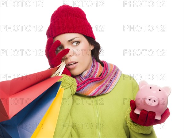 Concerned expressive mixed-race woman wearing winter clothing holding shopping bags and piggybank isolated on white background