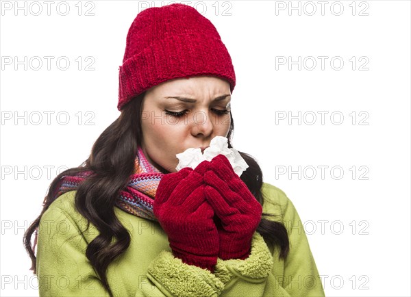 Sick mixed-race woman wearing winter hat and gloves blowing her sore nose with a tissue isolated on white background
