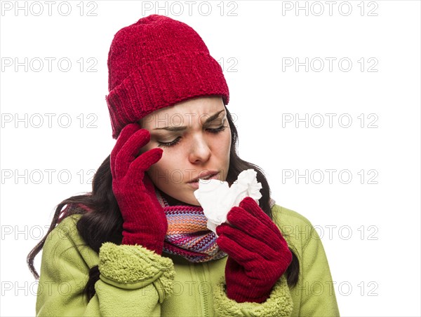 Sick mixed-race woman wearing winter hat and gloves blowing her sore nose with a tissue isolated on white background