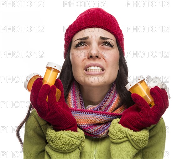 Sick mixed-race woman wearing winter hat and gloves blowing her sore nose and holding empty medicine bottle isolated on white