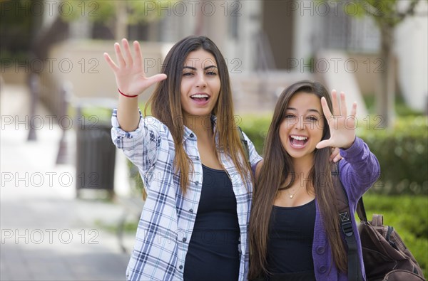 Portrait of two attractive mixed race female students waving and carrying backpacks on school campus