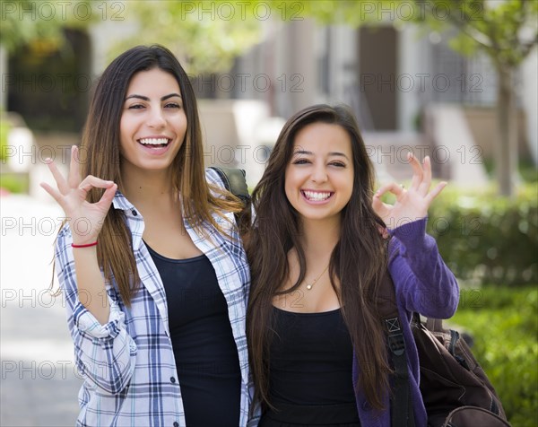 Portrait of two attractive mixed race female students with okay hand sign and carrying backpacks on school campus