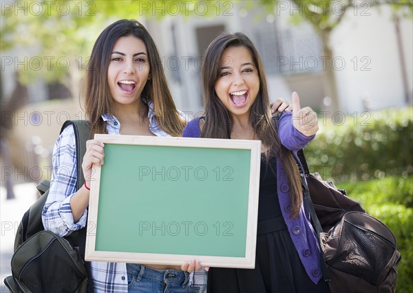 Portrait of two attractive mixed race female students holding blank chalkboard with thumbs up and carrying backpacks on school campus