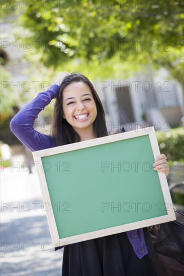 Portrait of an attractive excited mixed race female student holding blank chalkboard and carrying backpack on school campus