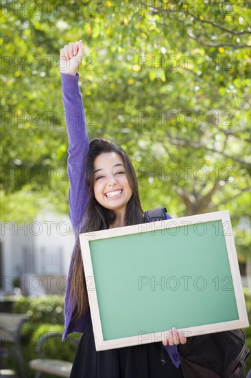 Portrait of an attractive excited mixed race female student holding blank chalkboard and carrying backpack on school campus