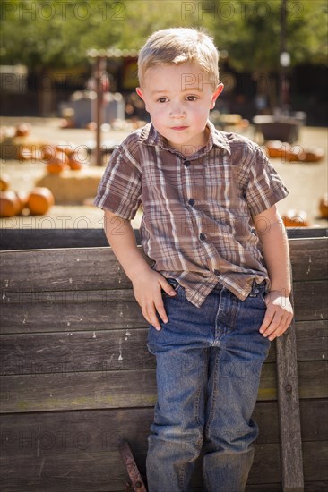 Adorable little boy at pumpkin patch with hands in his pockets leaning on antique wood wagon in rustic ranch setting