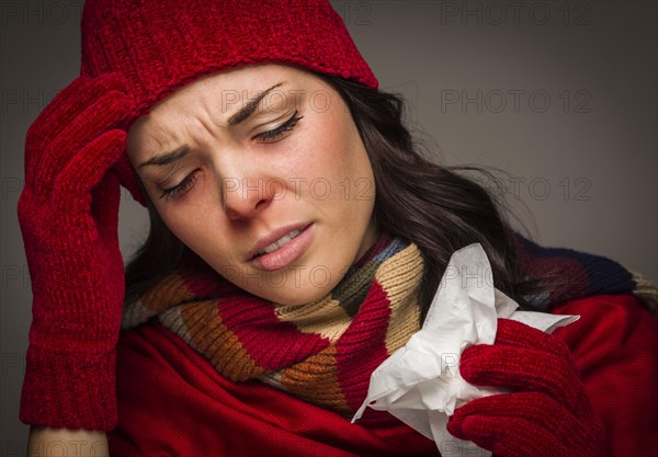 Sick mixed-race woman wearing winter hat and gloves blowing her sore nose with a tissue