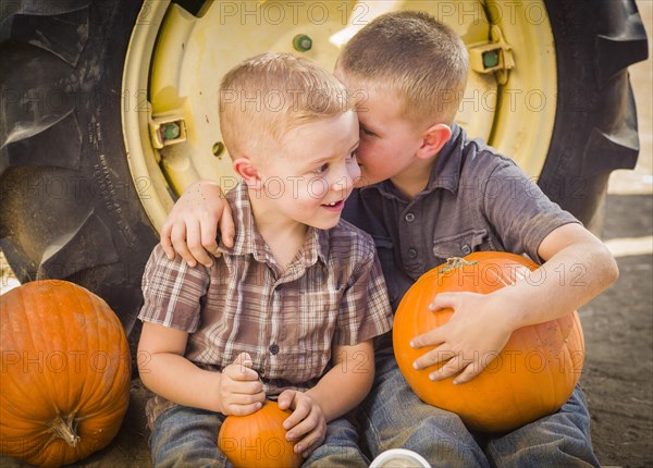 Two boys sitting against a tractor tire holding pumpkins and whispering secrets in rustic setting