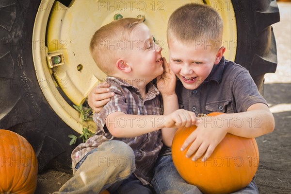 Two boys sitting against a tractor tire holding pumpkins and whispering secrets in rustic setting