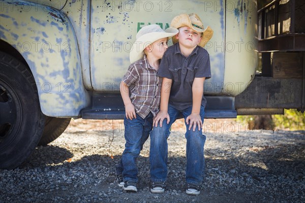 Two young boys wearing cowboy hats leaning against an antique truck in a rustic country setting