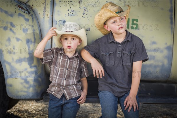 Two young boys wearing cowboy hats leaning against an antique truck in a rustic country setting