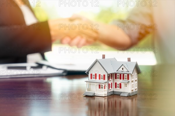 Real estate agent and customer sign contract papers and shake hands with small model home in front