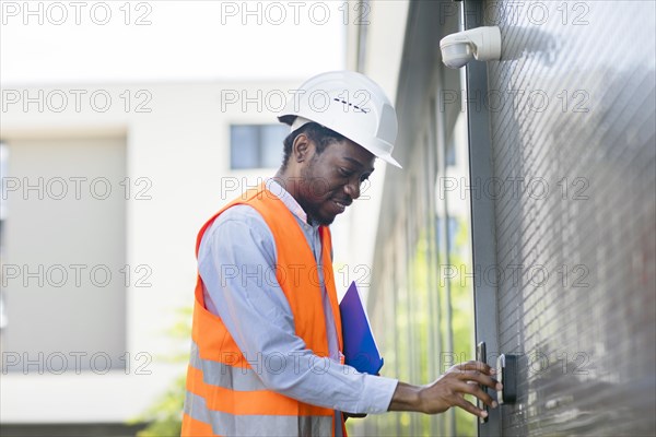 Young black man works outside as technician checks with smartphone and helmet and safety vest