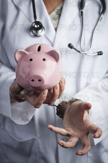 Female doctor or nurse in handcuffs holding piggy bank wearing lab coat and stethoscope
