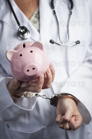 Female doctor or nurse in handcuffs holding piggy bank wearing lab coat and stethoscope