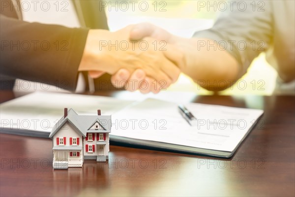 Real estate agent and customer sign contract papers and shake hands with small model home in front