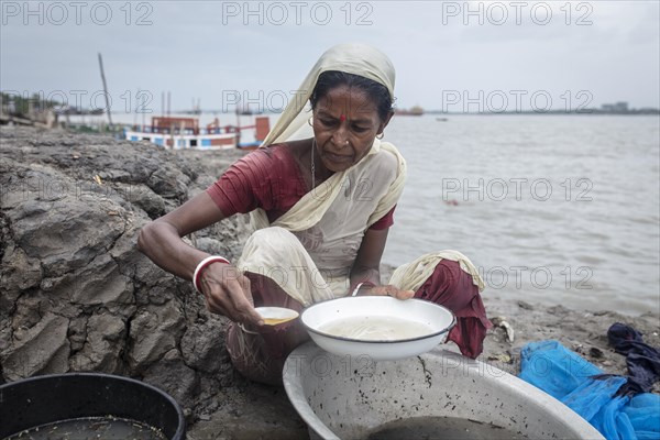 A woman sieves river water from wash bowls on a muddy embankment