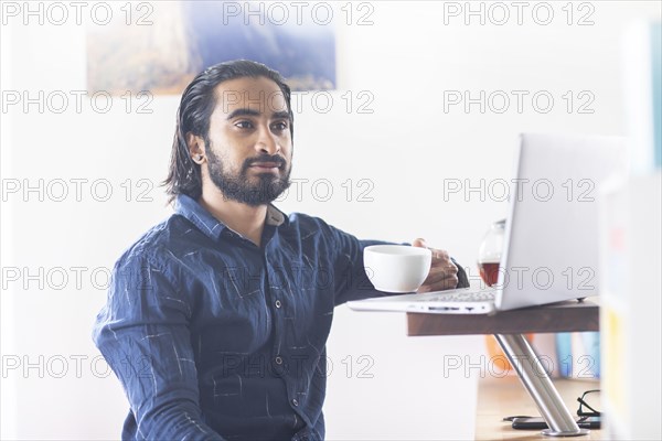 Young man with ponytail and checkered shirt drinking tea at home and working