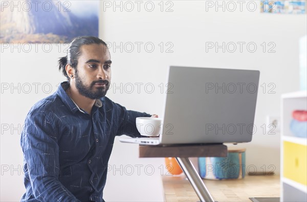 Young man with ponytail and checkered shirt drinking tea at home and working