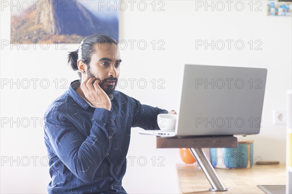Young man with ponytail and checkered shirt drinking tea at home and working