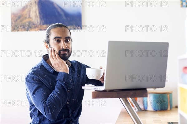 Young man with ponytail and checkered shirt drinking tea at home and working