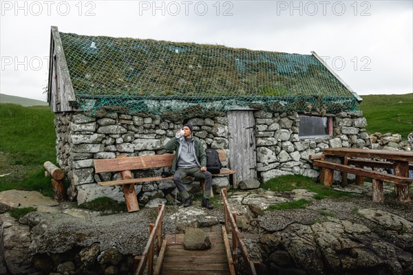 Hiker drinking water on a bench in front of a stone hut