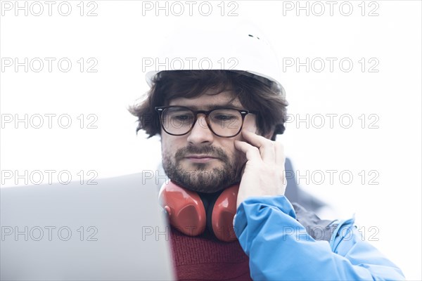 Young engineer with helmet and hearing protection checks outside work with laptop