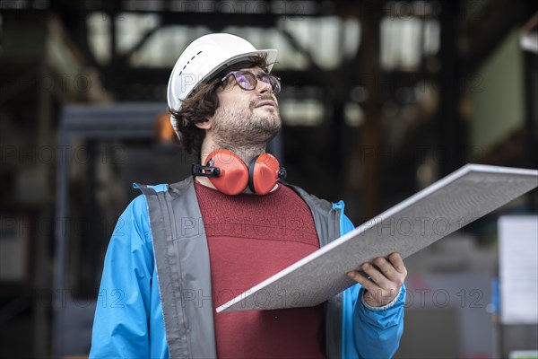 Young engineer with helmet and hearing protection checks with tile in front of a warehouse