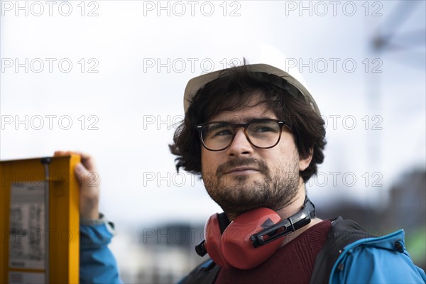 Young engineer with helmet and hearing protection checks outside work with laptop