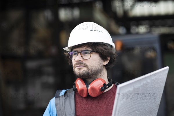 Young engineer with helmet and hearing protection checks with tile in front of a warehouse