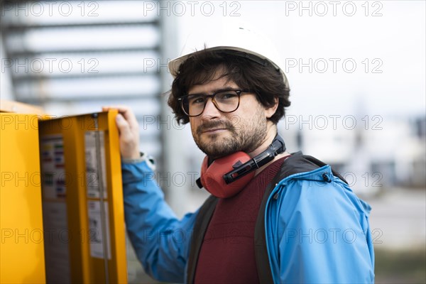 Young engineer with helmet and hearing protection checks outside work with laptop