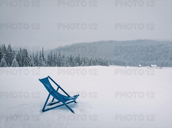 Sunbed type chair on the top of a mountain in a snowy winter day. Fir forest covered with snow on the horizon. Blizzard scene