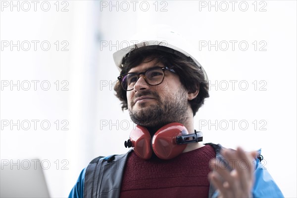 Young engineer with helmet and hearing protection checks outside work with laptop