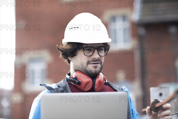 Young engineer with helmet and hearing protection checks outside work with laptop