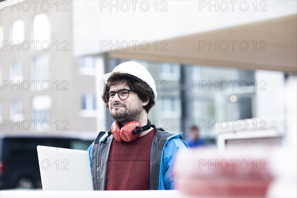 Young engineer with helmet and hearing protection checks outside work with laptop