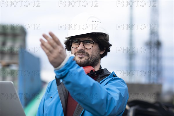 Young engineer wearing a helmet and hearing protection at a workplace outside with a laptop