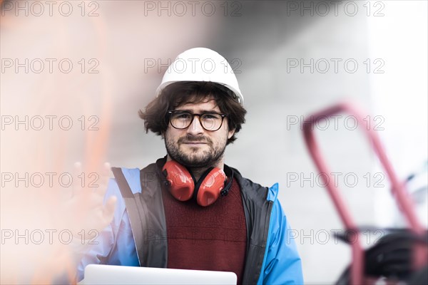 Young engineer with helmet and hearing protection checks outside work with laptop