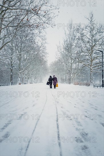 Two middle age women carrying pouches walks through the snowfall alongside the alley in the winter park. Wonderful snowing scene on the street. Cold season beauty. Blizzard in the Rose Valley city square