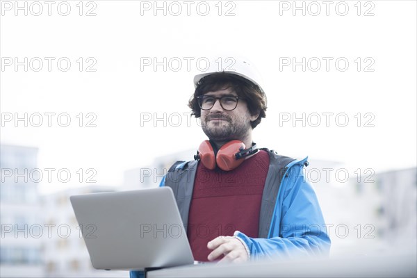 Young engineer with helmet and hearing protection checks outside work with laptop