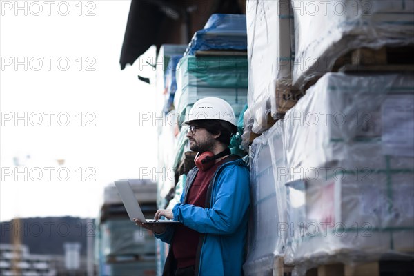 Young engineer with helmet and hearing protection checks outside work with laptop