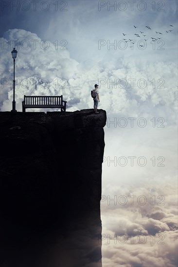 Inspirational imaginary view as a young boy with a bag in his back stand on the peak of a cliff above clouds