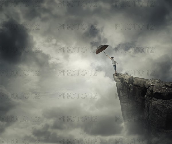 Boy trying to catch his umbrella on the edge of a cliff above the clouds