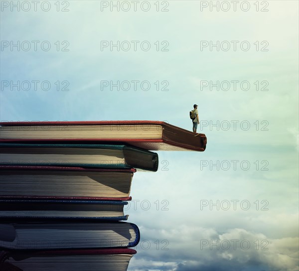 Young boy with a bag on his back stand on a stack of books looking far at horizon